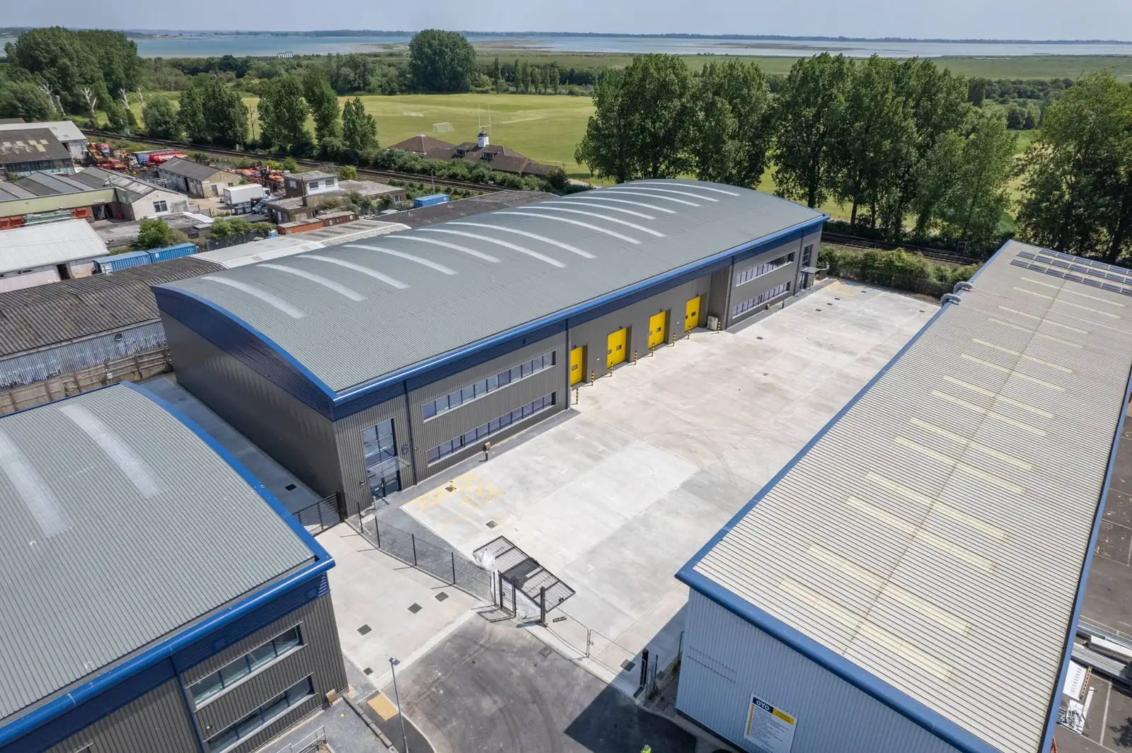 Aerial view of a modern industrial unit with a curved roof and yellow loading doors, with green fields and water beyond