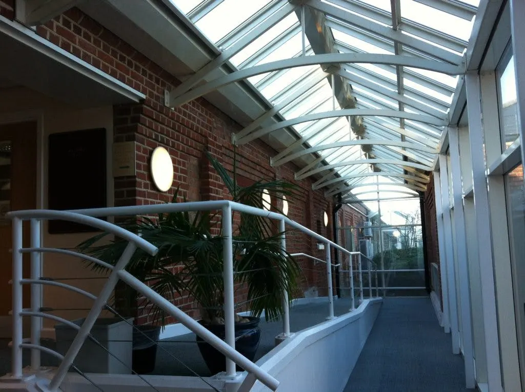 Glazed walkway with a white balustrade, plants and a glass roof beside a brick office building