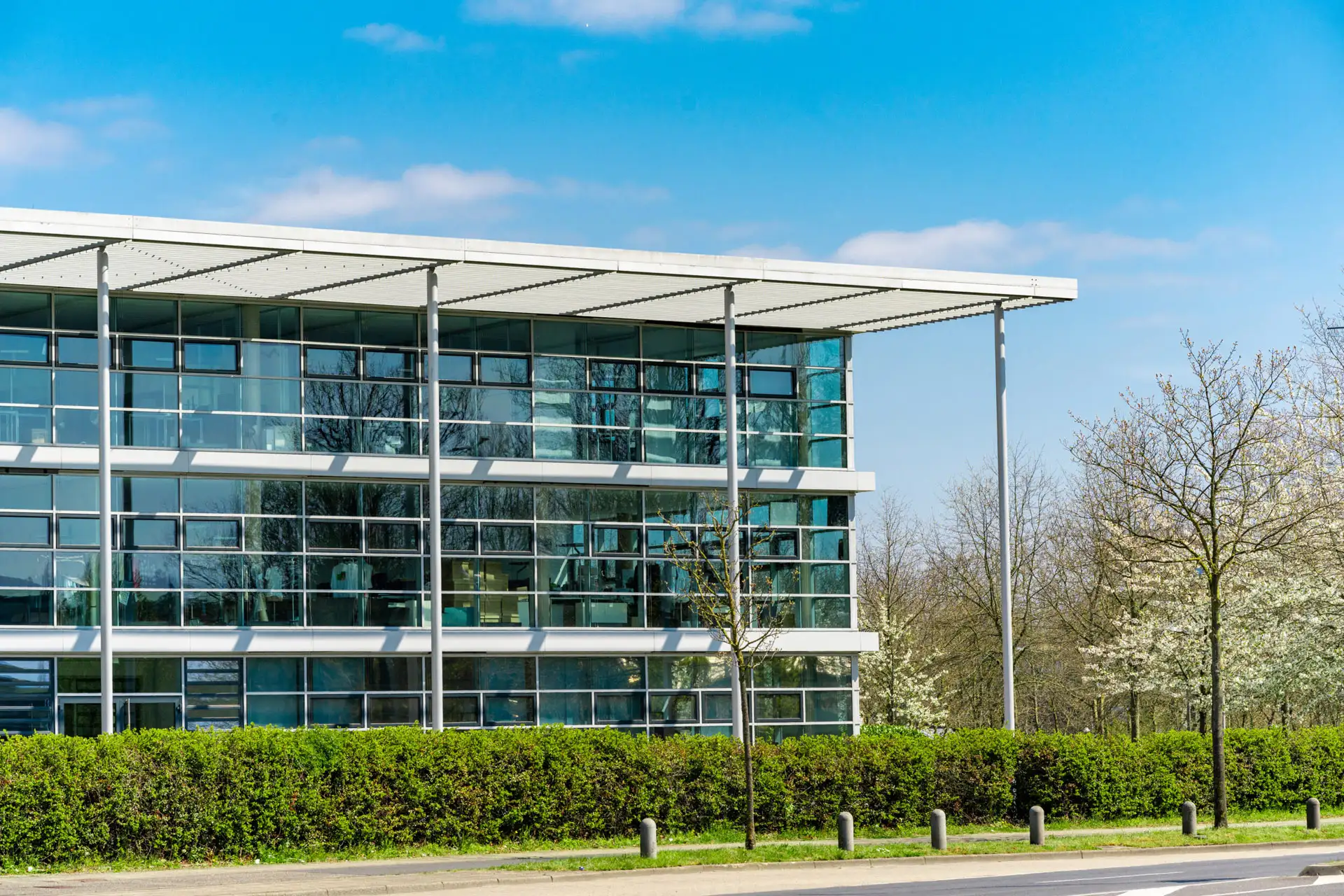 Glass-fronted office building with a flat canopy roof behind a clipped hedge