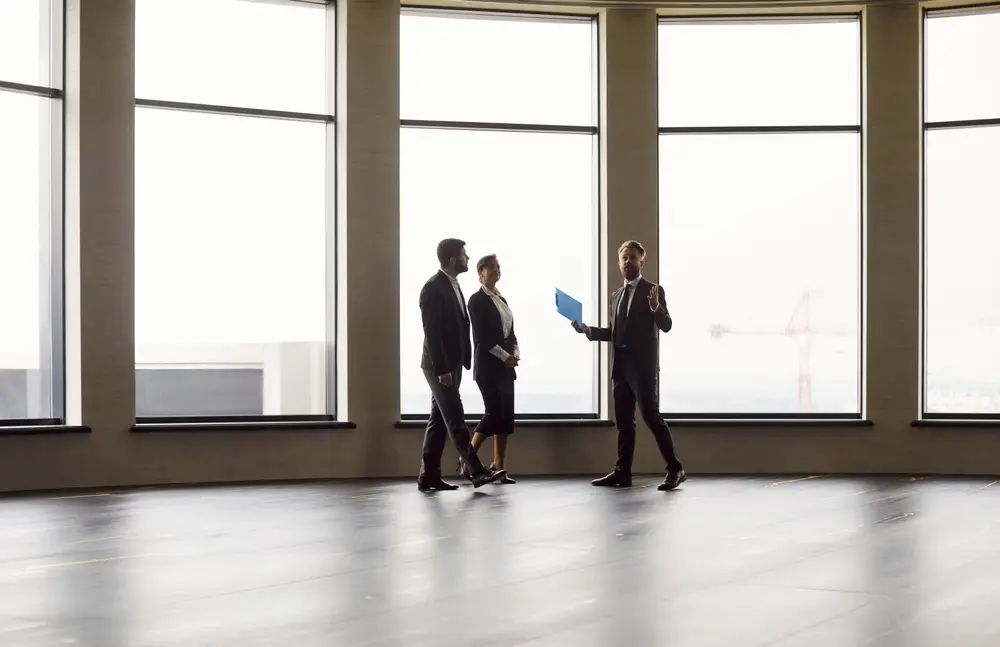Three business people standing in a bright empty office with tall windows, one holding plans