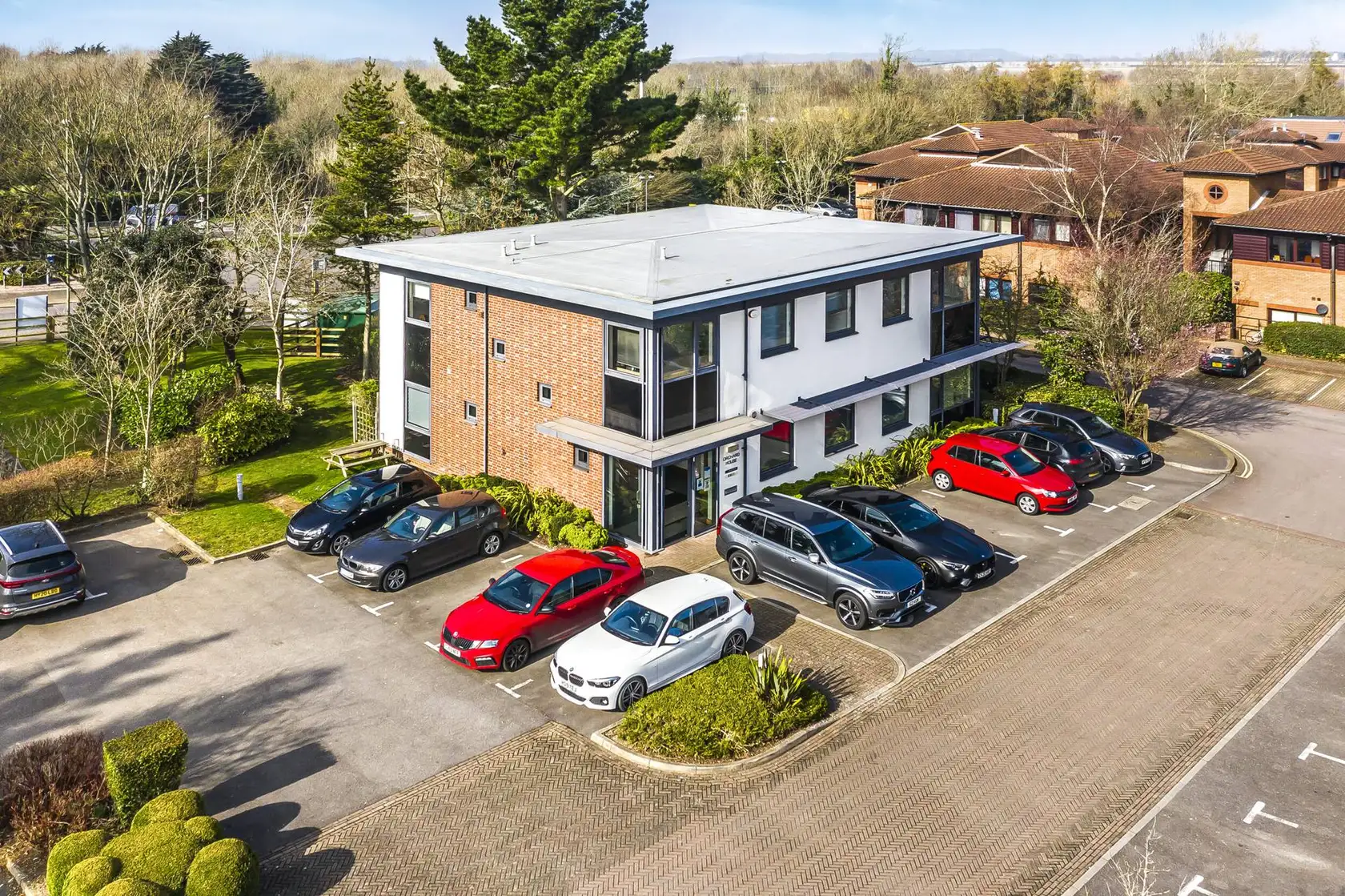 Aerial view of a brick office building with a car park and red and white cars beside trees